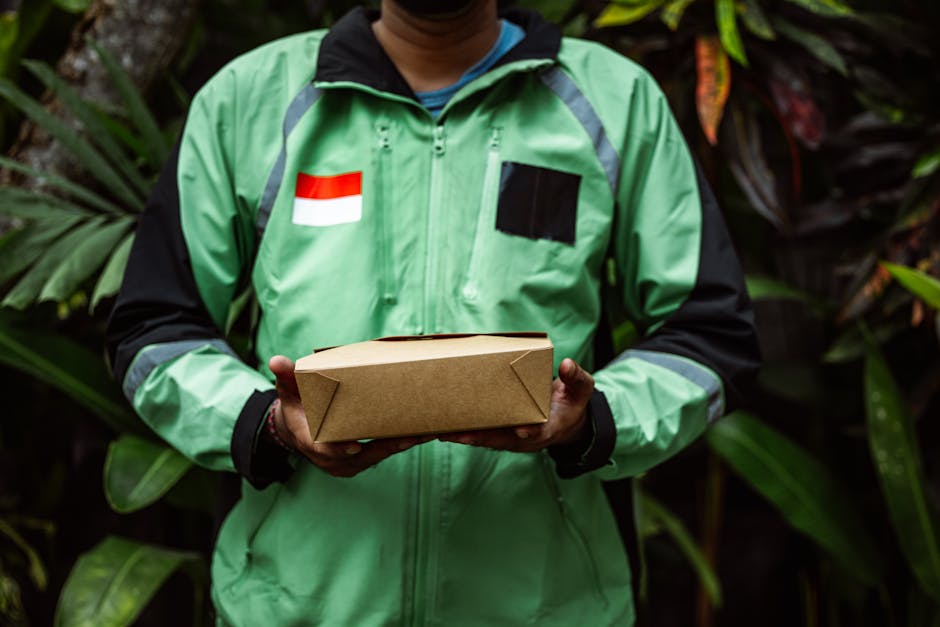 A delivery person wearing a bright green jacket with black accents, standing outdoors amidst lush green foliage, holding a rectangular brown paper package with both hands. The jacket features a red an